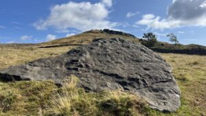 A large, flat earth-fast boulder of dark grey sandstone sits in the foreground, surrounded by dry moorland grass. Its weathered surface is pitted with dozens of small, circular cup-marks, a form of prehistoric rock art. In the background, the steep, grassy slope of Garfit Gap rises toward a blue sky with scattered white clouds, its summit capped by the jagged sandstone pillars and columns of The Wainstones. A low dry stone wall and two sparse trees are visible in the mid-ground.