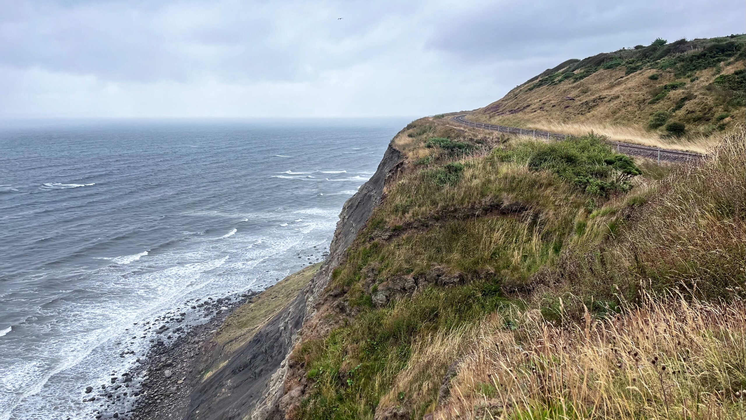 A single-track railway line of the East Cleveland Rail Line curves precariously along the rugged clifftop of Huntcliff, passing within 15 metres of the edge. To the left, the dark, sheer cliff face drops toward a choppy sea and a narrow rocky beach. Situated at the foot of these cliffs is a wide, lower vegetated platform known as Green Turf, a site identified as a historic landslide feature. To the right of the track, a steep cutting slope covered in tall, dry grass and patches of green scrub rises sharply upward. The scene is captured under a grey, overcast sky, emphasising the isolation and severe geotechnical risks of this coastal rail section.