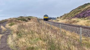 A heavy freight train, led by a blue and yellow locomotive hauling a long string of silver hopper wagons, rounds a sharp curve on the single-track East Cleveland Rail Line as it contours Warsett Hill above Huntcliff. The train travels on a bed of fresh grey ballast, passing a white "W" whistle board positioned at the side of the track. To the right, the steep hillside is covered in vibrant patches of purple heather and dry, wind-swept grass. To the left, a wire-and-post fence separates the line from a clifftop path that leads toward a large, circular metal sculpture visible on the horizon. The sculpture’s official name is Circle, but locals fondly know it as “the Charm bracelet”. The piece was created by Richard Farrington and is a familiar landmark on the Cleveland Way footpath. The scene is set under a flat, grey sky, capturing the train navigating the narrow corridor between the steep cutting of the hill and the nearby cliff edge.