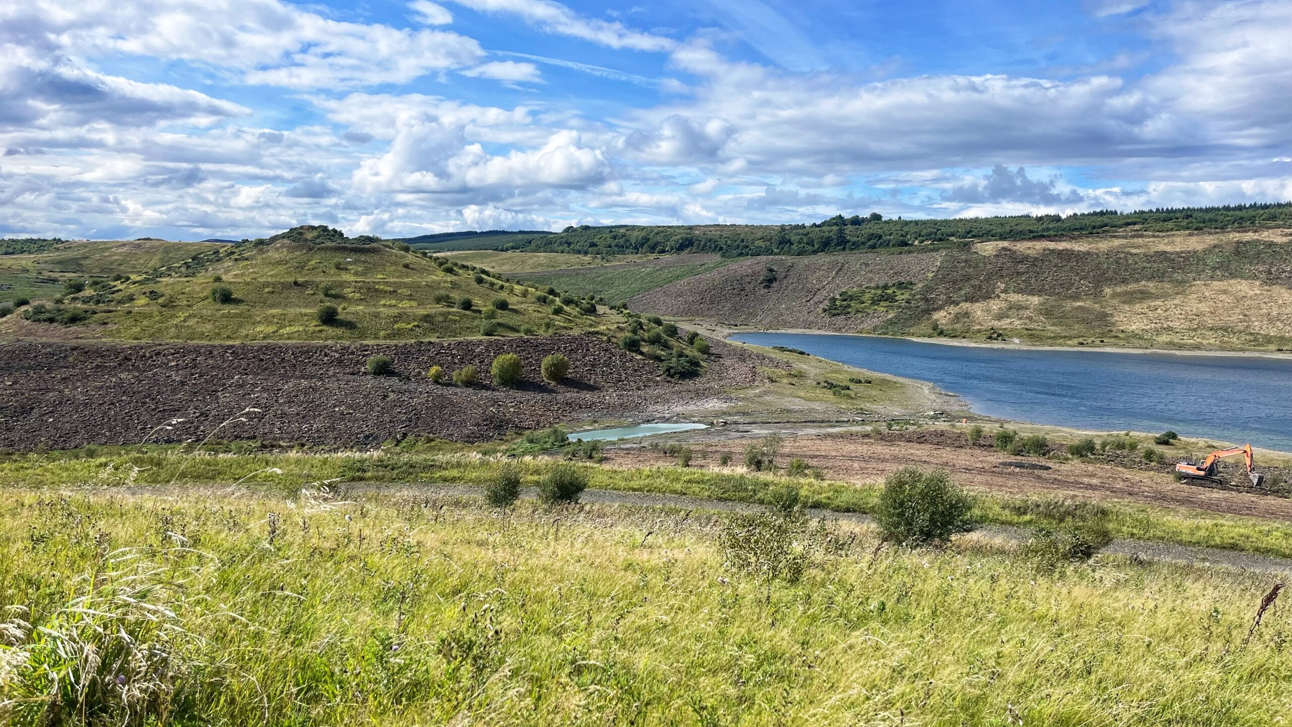 A manmade lake sits below rolling hills, the water a cool blue against banks of bare grey rock and rubble. Rough grassland climbs the slopes, dotted with young trees and scrub still finding their feet, and a dark line of forestry runs along the ridge behind. Down on the shoreline, near a small pool of pale turquoise water, an orange digger stands parked on a stretch of cleared track, a small reminder that this stretch of ground is still being coaxed back to life after its mining days.