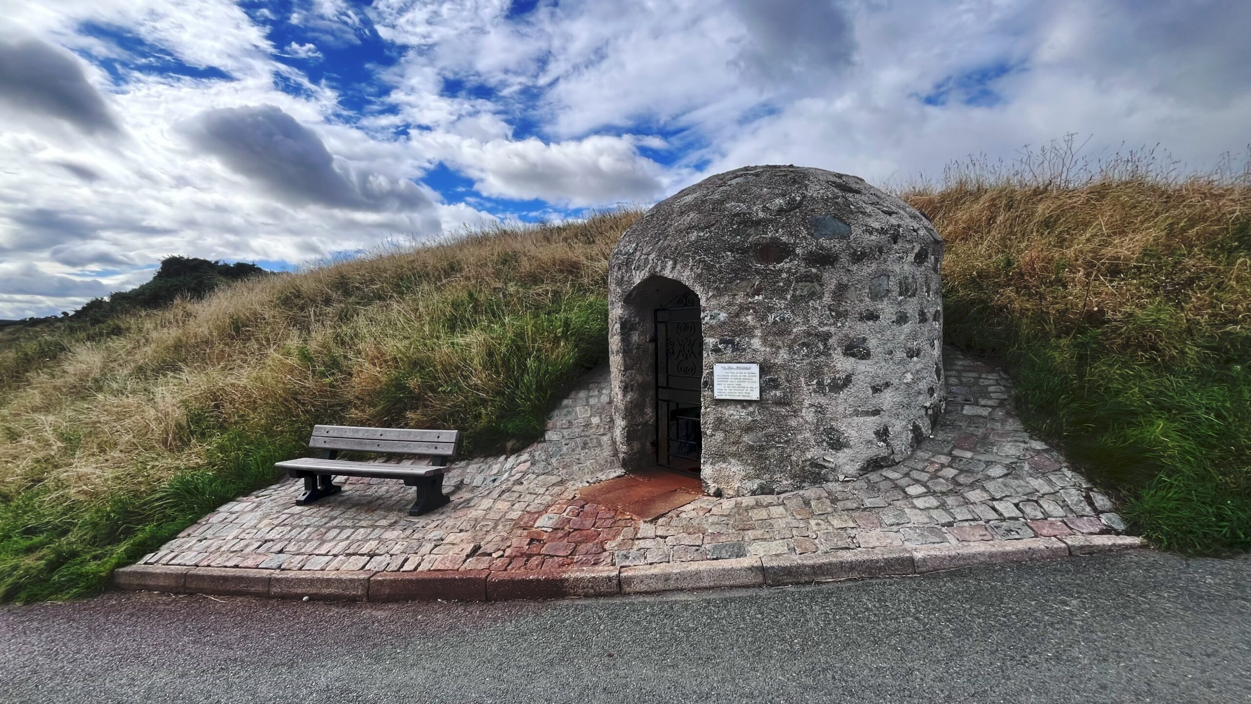 A small, dome-roofed stone well house, beehive-shaped and built from rough rubble, sitting at the bottom of a grassy bank beside a coastal path. A rounded stone archway holds an iron gate over the entrance, and a plaque is fixed to the wall beside it. On the cobbled ground at the threshold, a rusty red stain spreads out from the doorway, the mark left by the iron-rich spring water inside. A wooden bench sits to the left, and a paved path curves away to the right under a wide, cloud-streaked sky.