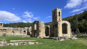 Ruins of Mount Grace Priory church in North Yorkshire under a bright blue sky with scattered white clouds. A square, battlemented stone tower with narrow pointed windows rises on the right, beside roofless walls that keep a large empty Gothic window. Low lines of foundation stones cross the neatly mown lawn in front, with a white wooden bench near the base of the walls. Longer ruined walls stretch away to the left, and a steep wooded hillside rises behind.