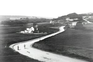 Black and white photograph looking northwest across the Esk valley at Castleton. A pale, winding road runs from the foreground into the distance, with two figures standing on it at lower left. A white cottage sits beside the road on the left. The surrounding fields and slopes are almost bare of trees. Bow Bridge is right of centre, with the Cleveland Inn and the railway behind it. A bare ridge fills the skyline.
