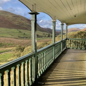 View along the open veranda of The Bungalow in Martindale, Lake District. Green wooden posts and balusters hold up a curved cream ceiling. Beyond the rail, oak trees in autumn colour sit below steep russet hills, and the balusters throw long shadows across the floor.