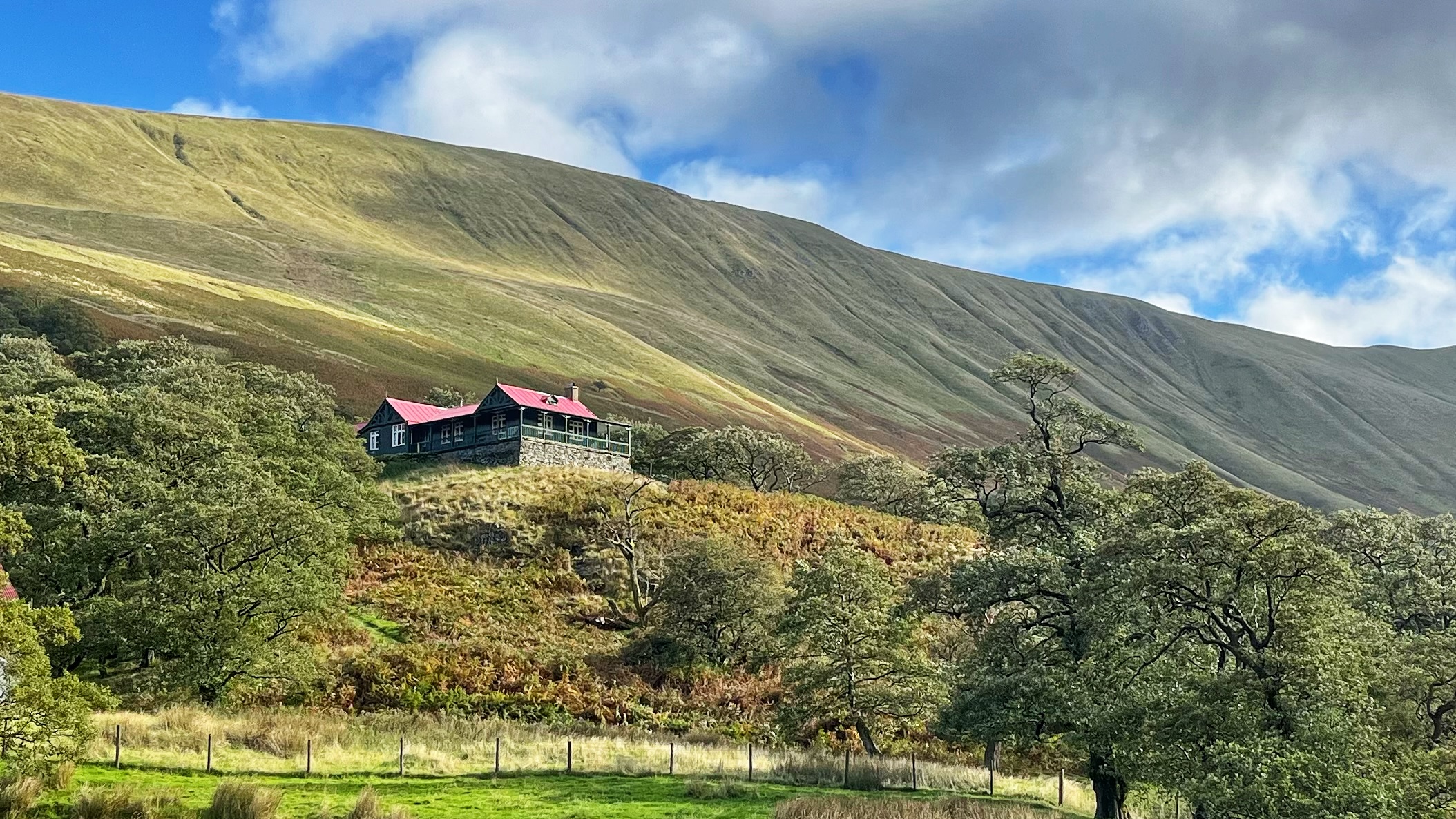 The Bungalow in Martindale, Lake District, a dark green wooden lodge with a red roof and a veranda, standing on a stone base halfway up a wooded hillside. A large grassy fell rises behind it under a blue sky with white clouds.