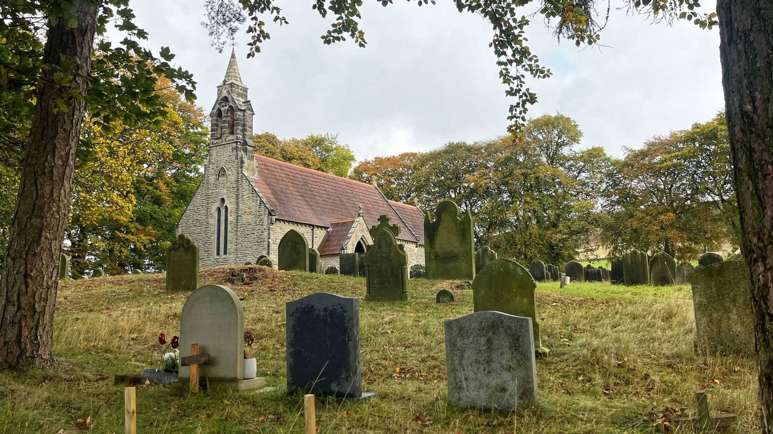 A historic stone church with a pointed bellcote and red-tiled roof stands on a hill surrounded by autumn trees, set behind a grassy churchyard filled with weathered gravestones.