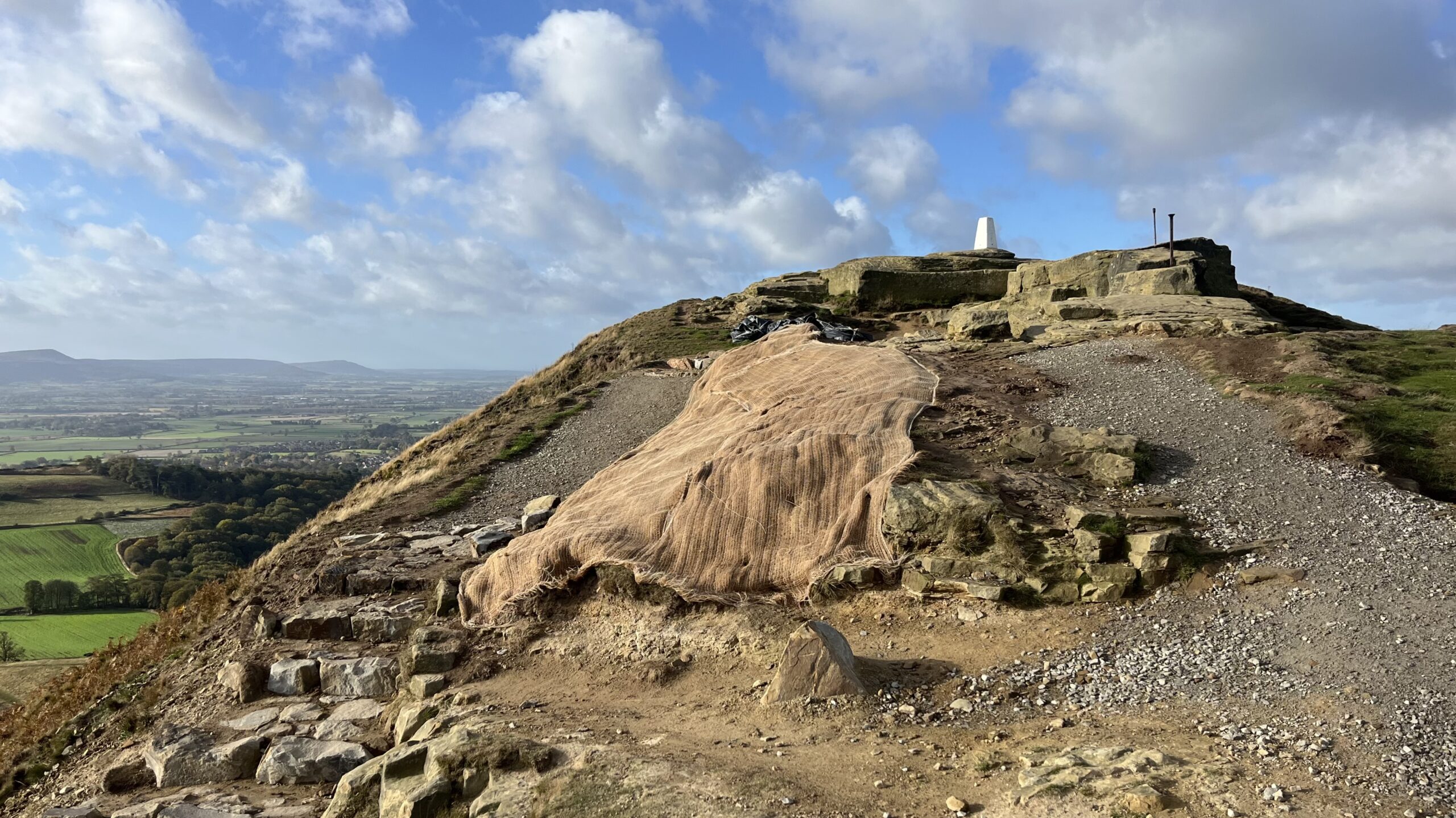 A rocky hilltop with a white trig point overlooking a broad green valley. A large section of exposed earth is covered with protective matting, surrounded by uneven stone steps, loose gravel and weathered rock. Patchwork fields, wooded areas and distant hills stretch beneath a blue sky filled with scattered white clouds.