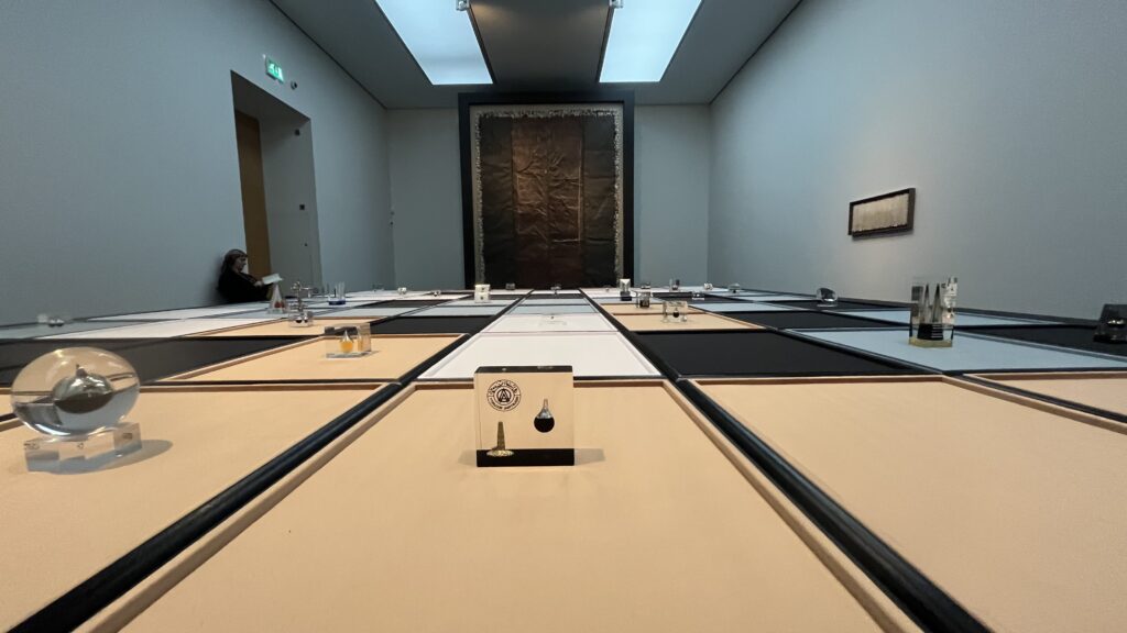 A low view across a grey gallery floor laid out as a grid of felt trays in peach, black, white and pale blue. Each tray holds a small clear paperweight with a dark droplet sealed inside. A woman sits reading in the left corner, and a large dark brown panel with a fringed edge hangs on the far wall.