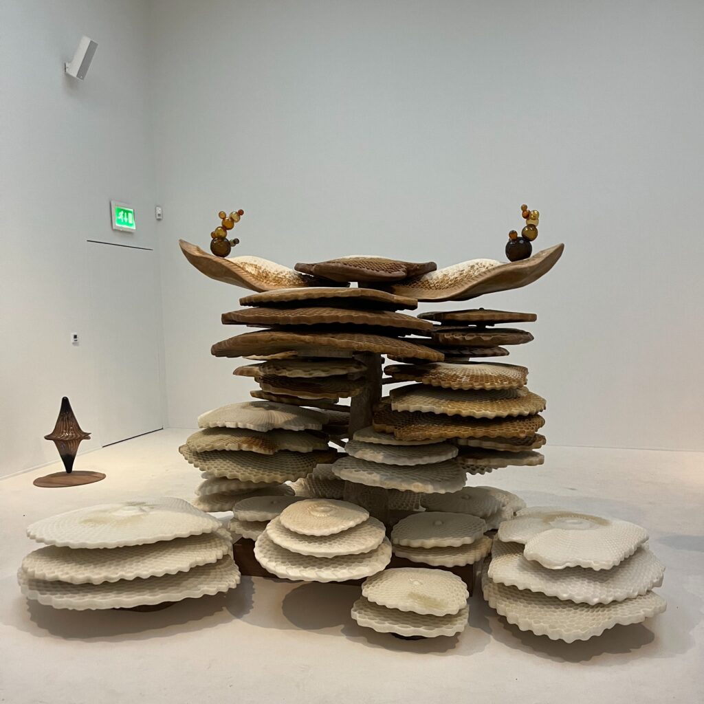 A tall sculpture made of stacked honeycomb discs in a white gallery. The discs are pale cream at the base and darken to brown at the top, where two clusters of amber glass beads sit on curved wooden forms. A small brown spinning-top shape stands on a disc to the left.