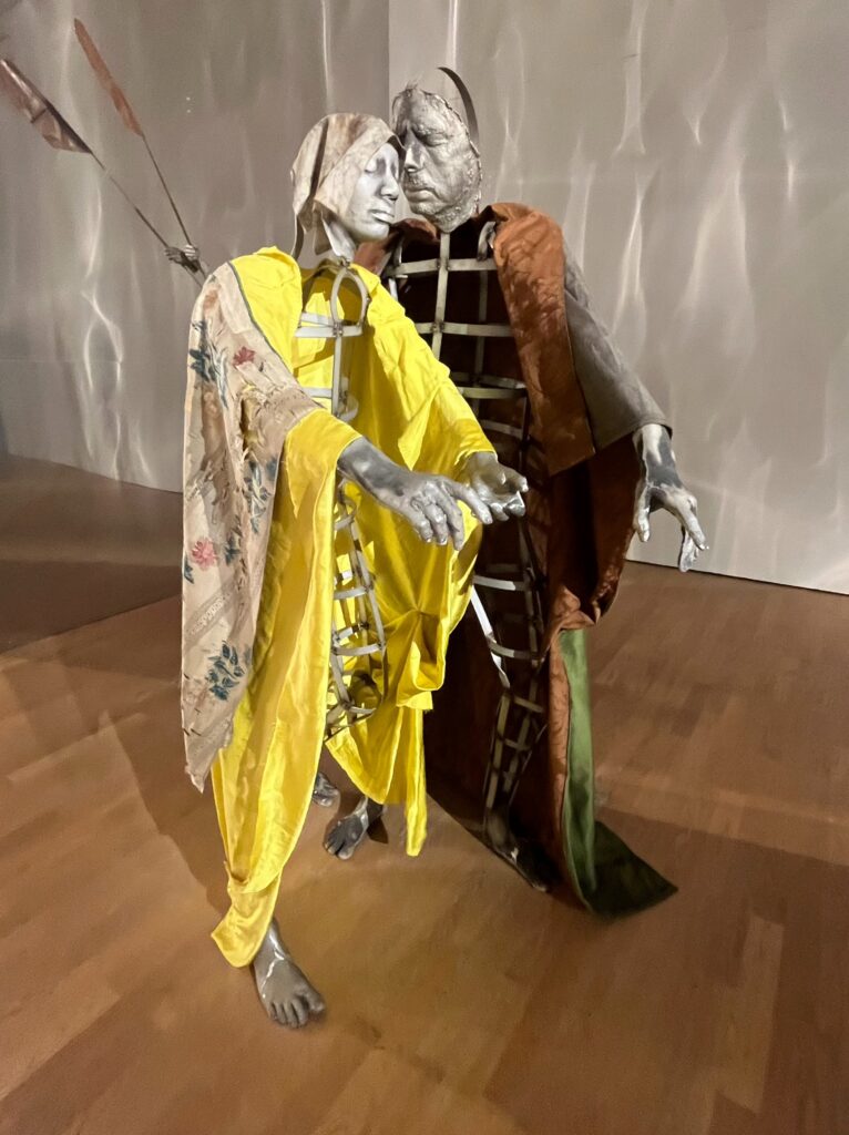 Two metal-ribbed figures with silver-grey faces lean their heads together. One wears bright yellow silk with a faded floral sleeve, and the other a copper-brown cloak lined in green. Both reach out with open hands and bare feet, and a pair of long spears or flags stands behind them.