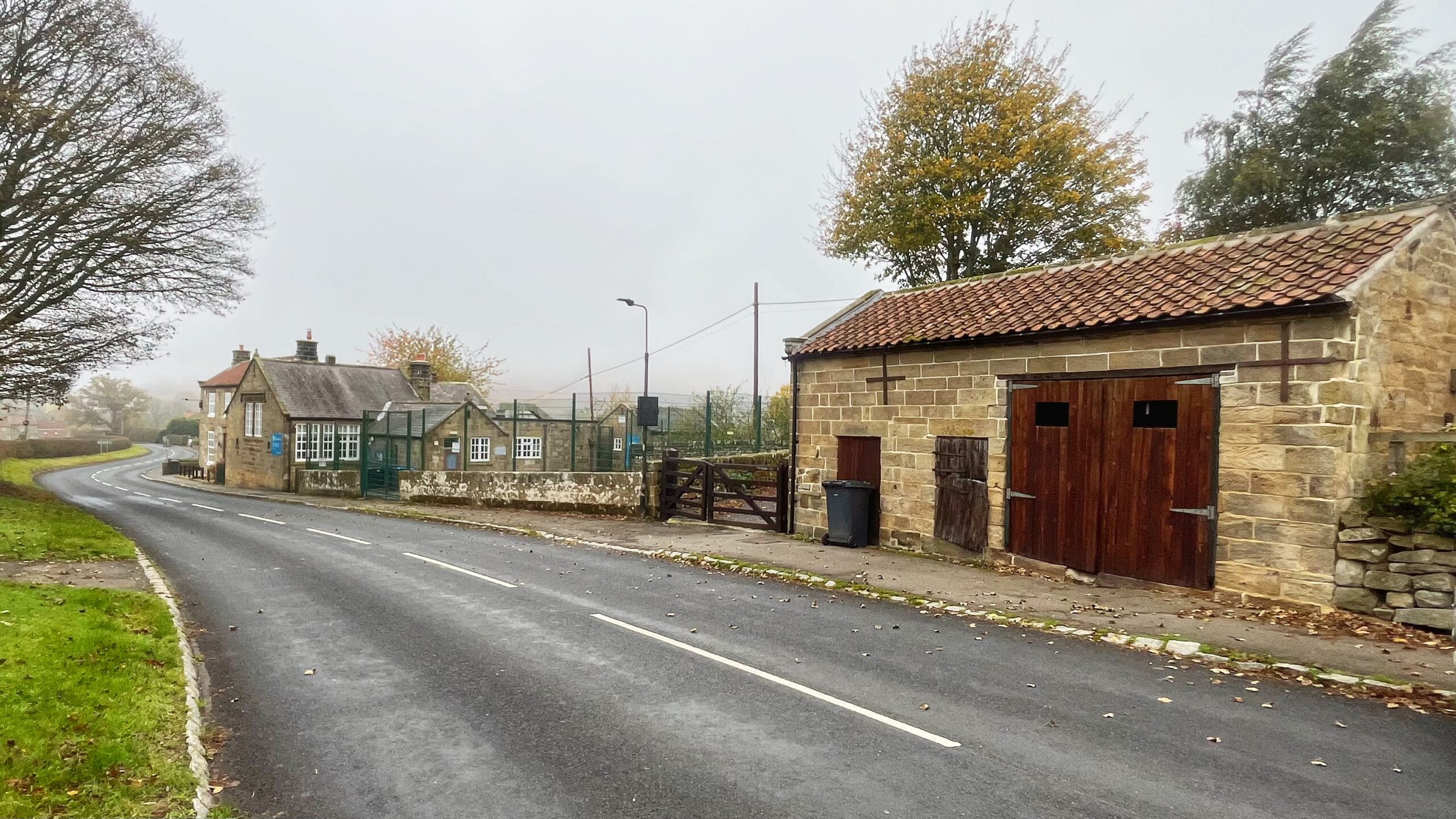 A quiet asphalt road curving through the overcast, misty village of Chop Gate. On the right stands a traditional stone outbuilding with a red pantile roof and dark wooden doors. Further down the road on the left sits Bilsdale Midcable CE Primary School, a single-story stone building with white-framed windows and a high, green fence surrounding the playground, framed by autumn trees.